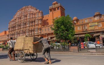 Zwei Händler transportieren ihre Waren am Hawa Mahal, auch Palast der Winde genannt, in Jaipur vorbei. Das fünfstöckige Gebäude hat 953 Jharokhas (kleine Fenster mit komplizierten Gitterverstrebungen), von denen aus die königlichen Damen das Treiben auf der Straße beobachten konnten, ohne gesehen zu werden.