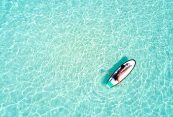 Das Meerwasser der Malediven ist berühmt für seine wunderschöne aquamarinblaue Farbe. Auf dieser Luftaufnahme ist eine Frau zu sehen, die sich auf einem Surfbrett entspannt © Sven Hansche