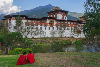 Zwei angehende Mönche sitzen mit Blick auf den Punakha Dzong, Bhutan. Diese ist die zweitgrößte und zweitälteste Klosterfestung des Königreichs und steht anschaulich für die bhutanische Handwerkskunst und Architektur von Weltklasse