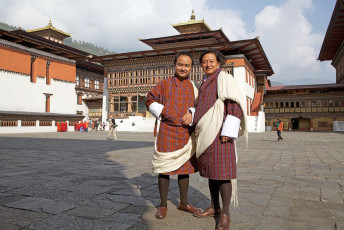Bhutans Nationaltracht für Männer, der Gho, geht auf das 17. Jahrhundert zurück und besteht aus einem knielangen Gewand, das in der Taille mit einem handgewebten Gürtel, dem Kera, gebunden wird. Zu besonderen Anlässen wird ein weißer Seidenschal, der sogenannte Kabney, getragen. Auf dem Bild posieren zwei Männer für die Kamera am Trashi Chho Dzong in Thimphu, Bhutan © Antonella865
