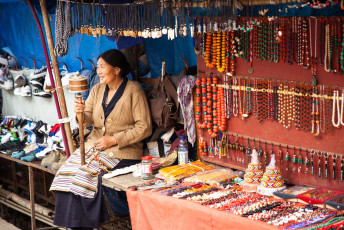Eine tibetische Frau neben ihrer Schmuckauslage auf einem Straßenmarkt in Dharamsala. In ihrer Hand hält sie eine traditionelle Gebetsmühle © Elena Ermakova