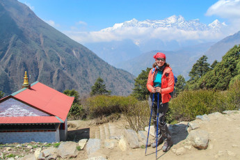 Eine Bergsteigerin mit ihrem Tagesrucksack lächelt in die Kamera, nachdem sie am Eingang des Dorfes Thyangboche im Himalaya-Gebirge in Nepal angekommen ist. Im Hintergrund ist dar Kongde Ri-Gipfel zu sehen, einer der schwieriger zu besteigenden Berge. © Andrey Rykov