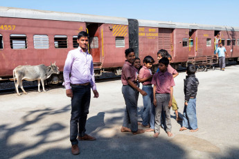 Eine Gruppe von Schuljungen in Uniform lächelt am kleinen Bahnhof von Phulad in die Kamera, während im Hintergrund der Zug aus Khamlighat ankommt. Eine heilige Kuh schaut zu © Bim