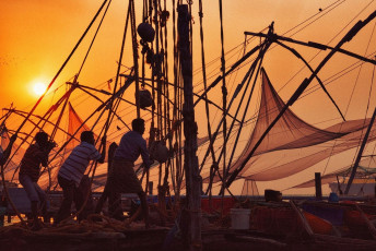 Eine Gruppe von Männern reiht sich auf, um chinesische Fischernetze einzuholen, während die Sonne in Kochi, Kerala, untergeht. Chinesische Fischernetze wurden vor über 500 Jahren bei den Fischern in Kochi eingeführt © powerofforeever